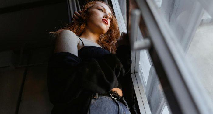 A woman gazes out of a window, deep in thought in a softly lit room.