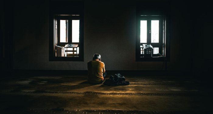 A solitary figure sits in contemplative silence in a dimly lit room with soft natural light.