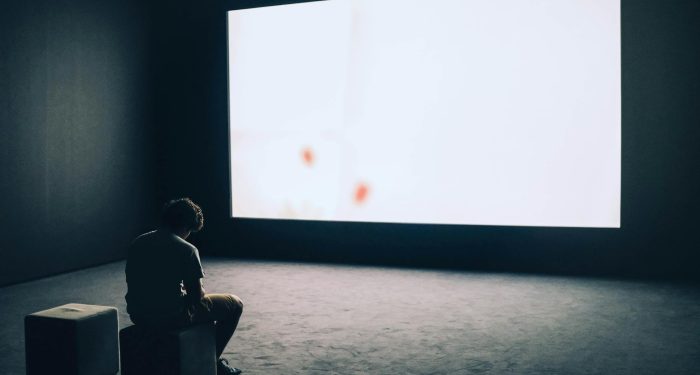 A man sits alone in a dark room with a large white screen, depicting solitude and contemplation.