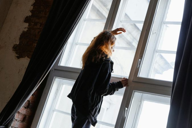 A woman stands in deep thought near a large window, embracing solitude.