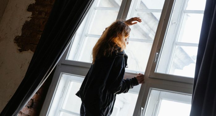A woman stands in deep thought near a large window, embracing solitude.