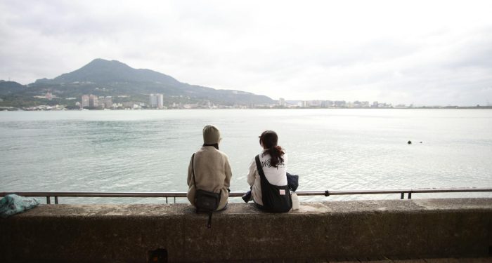 Two people sitting by the waterfront, gazing at distant mountains.