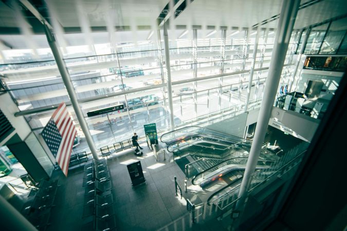 A bright, spacious airport terminal featuring expansive glass walls, escalators, and American flag.