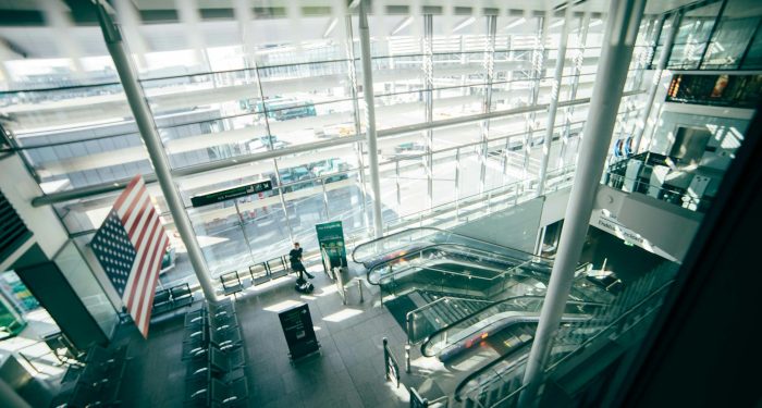 A bright, spacious airport terminal featuring expansive glass walls, escalators, and American flag.