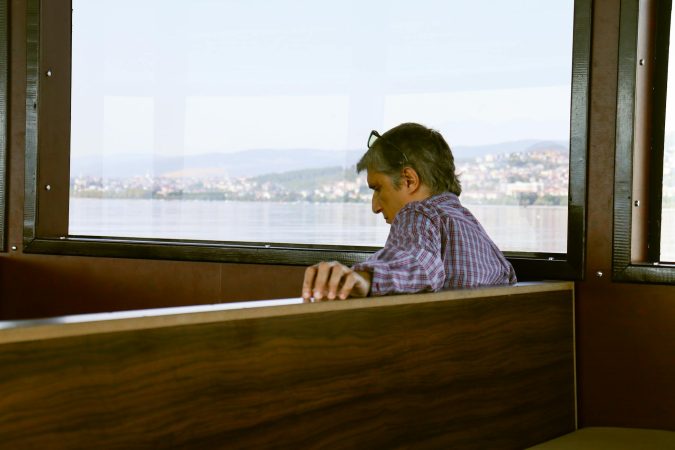 A man sits alone on a boat, gazing at serene waters with a distant city view.