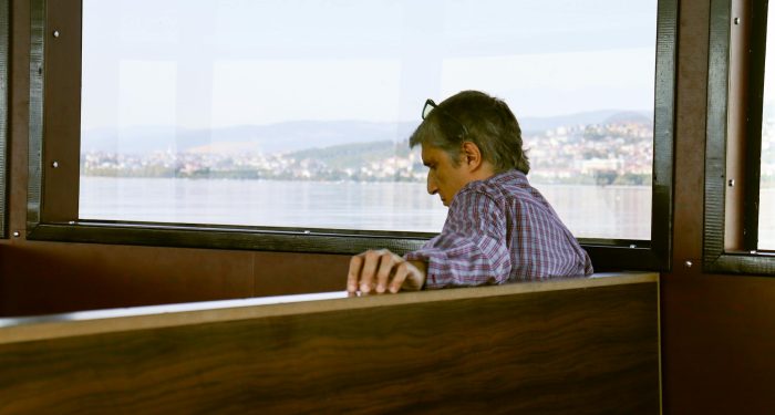 A man sits alone on a boat, gazing at serene waters with a distant city view.