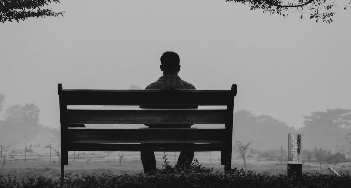 A solitary man sits on a bench in Dhaka, Bangladesh, silhouetted against a foggy, grayscale landscape.