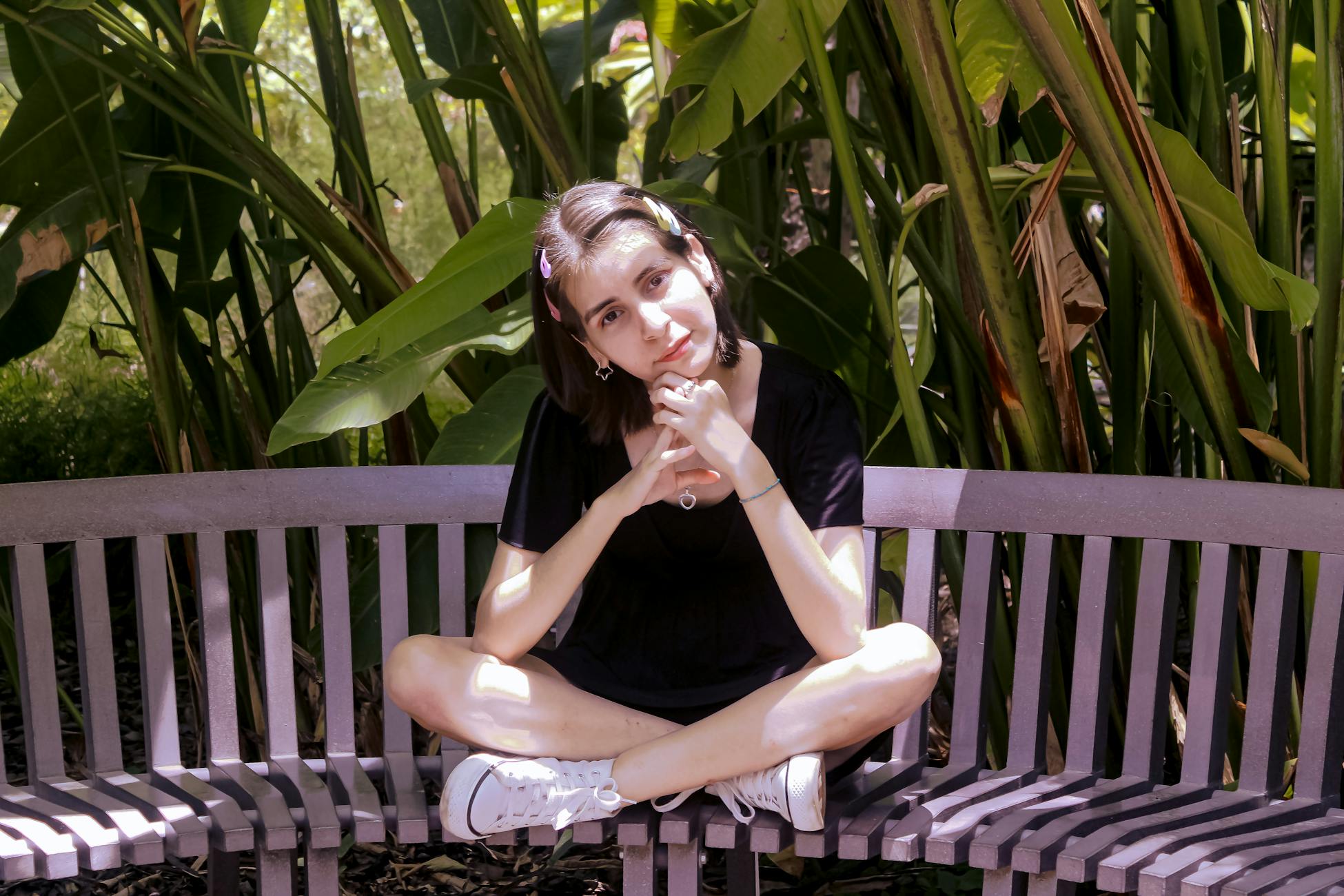 A young woman sits cross-legged on a bench surrounded by lush green plants, soaking up the natural ambiance.
