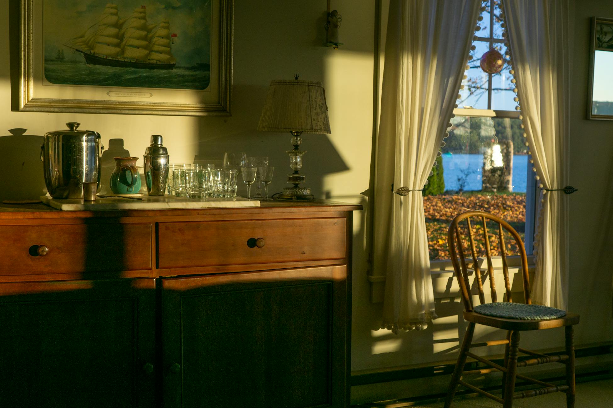 Warm interior scene with a wooden sideboard and sunlit chair near a window.