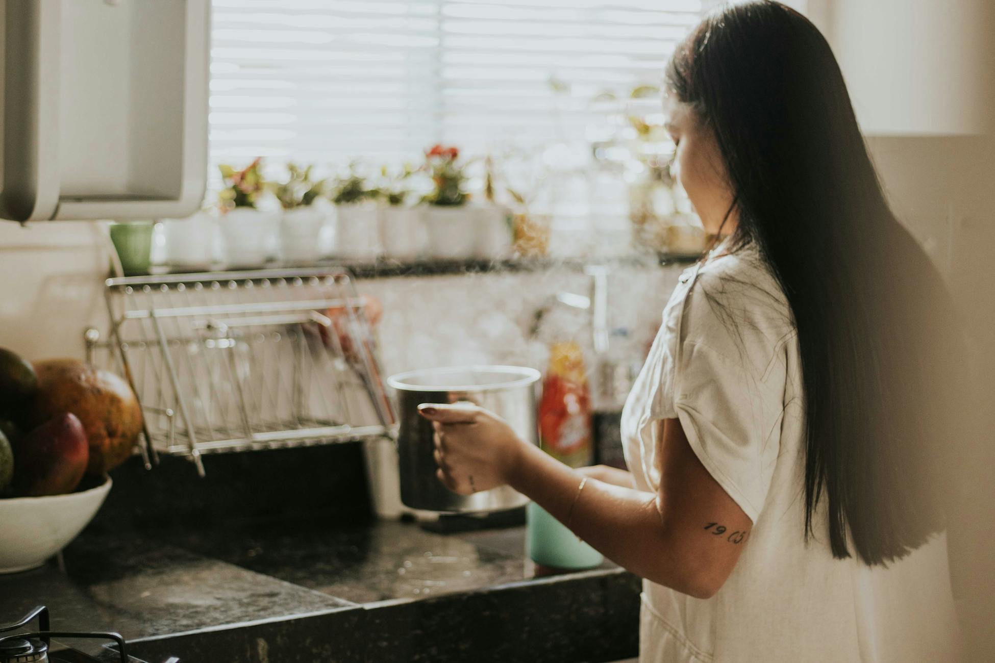 A woman cooking on a stovetop in a cozy, sunlit kitchen with houseplants on the windowsill.