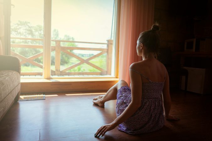 A woman sits inside a sunlit room looking out a large window, enjoying nature.