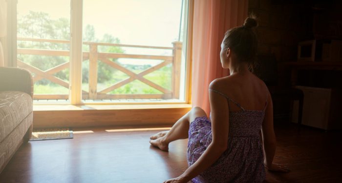 A woman sits inside a sunlit room looking out a large window, enjoying nature.