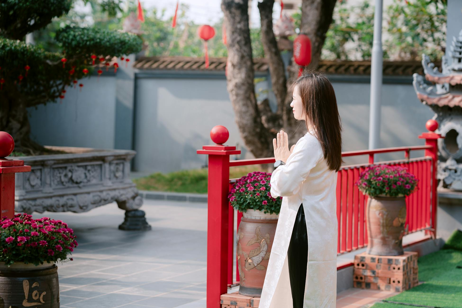 A woman meditates outdoors in a traditional Asian courtyard surrounded by vibrant flowers and red decorations.